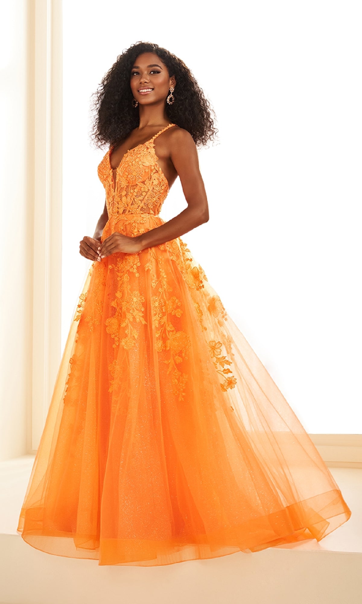 A woman poses indoors against a bright background, wearing the Ellie Wilde EW36015 long prom dress—a sleeveless, floor-length orange gown featuring lace embroidery and a tulle skirt.